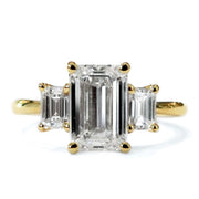A large emerald-cut diamond flanked by two smaller emerald cut diamonds form a classic three-stone engagement ring design set in yellow gold. Dana Walden NYC.